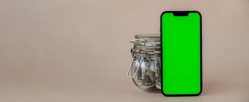 Banner Vertical Green screen on modern mobile phone in background of glass jar full of American currency dollar banknotes on beige background. Cope space for text. Advertisement for application website. Concept of money economy banks and finances photo