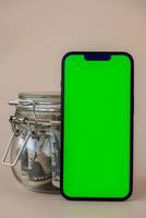 Chrome key smartphone screen mock up template in vertical position on beige background. Copy space App website advertising. Jar filled with dollars cash. Concept of Mobile application and technology business savings. photo