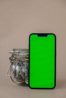 vertical verde pantalla en moderno móvil teléfono en antecedentes de vaso tarro lleno de americano moneda dólar billetes en beige antecedentes. capa pluvial espacio para texto. anuncio para solicitud sitio web. concepto de dinero economía bancos y finanzas foto