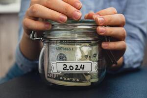 Unrecognizable woman holding Saving Money In Glass Jar filled with Dollars banknotes. 2024 transcription in front of jar. Managing personal finances extra income for future insecurity background photo