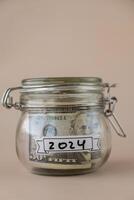 Saving Money In Glass Jar filled with Dollars banknotes. 2024 year transcription in front of jar. Managing personal finances extra income for future insecurity. Beige background photo