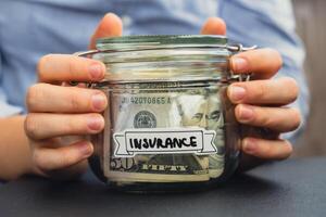 Unrecognizable woman holding Saving Money In Glass Jar filled with Dollars banknotes. INSURANCE transcription in front of jar. Managing personal finances extra income for future insecurity background photo