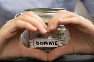 Female showing heart sign with hands Saving Money In Glass Jar filled with Dollars banknotes. DONATE transcription in front of jar. Managing personal finances extra income for future insecurity background photo