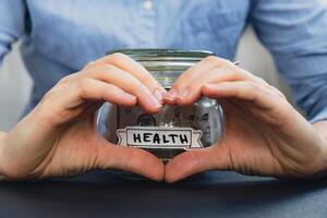 Female showing heart with hands sign Saving Money In Glass Jar filled with Dollars banknotes. HEALTH transcription in front of jar. Managing personal finances extra income for future insecurity background photo
