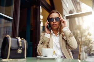 hermosa mujer vistiendo Gafas de sol sentado a café terraza hablando en teléfono mientras Bebiendo café foto