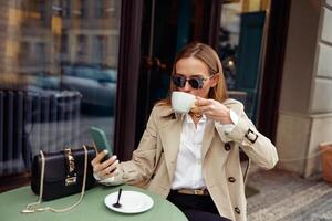 sonriente elegante mujer en lentes sentado a café terraza y utilizar teléfono mientras Bebiendo café foto