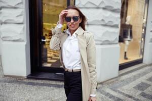 Happy fashionable woman in sunglasses standing on city street background and looking at camera photo