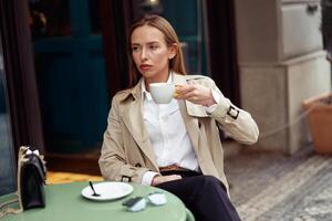 Stylish focused woman drinking coffee on restaurant terrace during break and looking at side photo