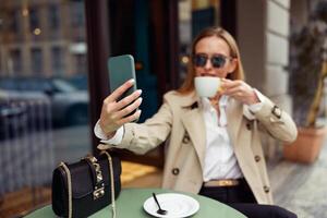 elegante mujer en lentes sentado a café terraza y tomando selfie en teléfono mientras Bebiendo café foto