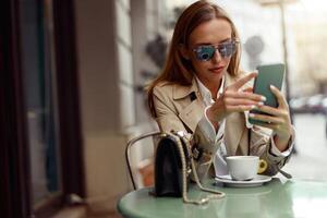 sonriente elegante mujer en lentes sentado a café terraza y participación teléfono mientras Bebiendo café foto