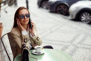 hermosa mujer vistiendo Gafas de sol sentado a café terraza hablando en teléfono mientras Bebiendo café foto