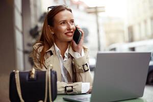 sonriente europeo mujer hablando teléfono mientras trabajando en línea sentado a al aire libre café terraza foto