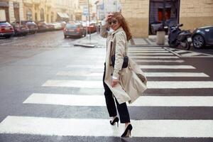 Woman in trendy outfits crosses road against backdrop of city buildings and looking at camera photo