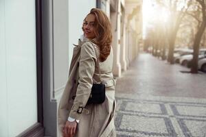 sonriente mujer en Saco cruce el ciudad calle y mirando a cámara. borroso antecedentes foto