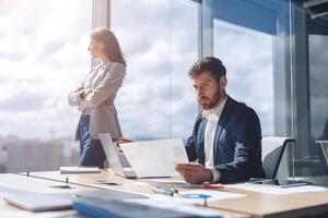 businessman analyzes work papers and charts while his colleague looks thoughtfully out the window photo