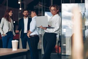 Mature confident businesswoman at office with group of colleagues on background, working on laptop photo