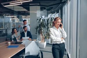 Focused businesswoman is standing near window and talking phone during meeting in modern office photo