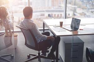 Focused businessman working on laptop and looking out the window at her workplace at modern office photo