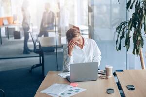 cansado mujer de negocios con dolor de cabeza trabajando en ordenador portátil a su lugar de trabajo a moderno oficina foto