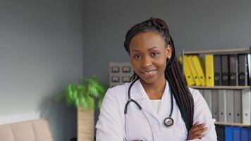 Portrait of handsome african female doctor wearing white coat and stethoscope crosses arms, standing in his health clinic office video