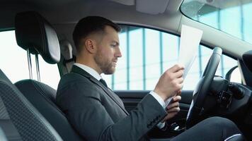 portrait de une sur de soi homme séance dans une voiture avec les documents dans une affaires costume près une moderne Bureau bâtiment video