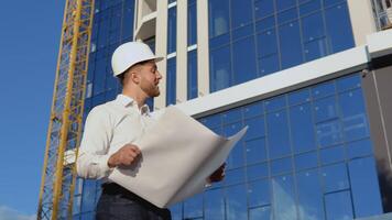 architetto ingegnere legge progetto disegni. un ingegnere nel un' bianca camicia e casco lavori su il costruzione di un' moderno bicchiere edificio video