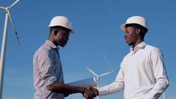 Two male African American electrical engineers stand against the backdrop of a windmill at an air power plant and shake hands video