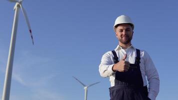 The power plant engineer is standing in a white classic shirt, blue overalls and a white helmet on a windmill background video