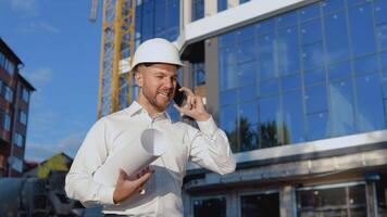 Evil engineer in a white shirt and helmet works on the construction of a modern glass building. The engineer-architect holds a roll with a drawing of the project and talks on a cell phone video