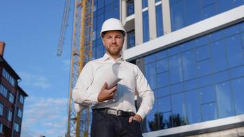 een ingenieur in een wit overhemd en helm staat tegen de achtergrond van een modern glas gebouw en houdt een rollen met een tekening van de project. modern bouw video