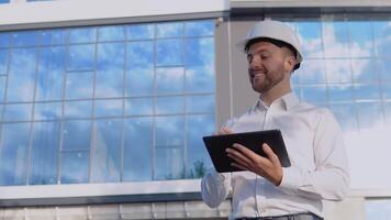 een ingenieur architect in een wit overhemd en helm Aan de achtergrond van een modern glas gebouw werken Holding een tablet en beheert de werk werkwijze video