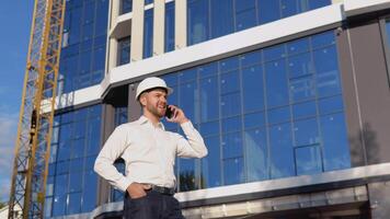 Engineer architect in a white shirt and helmet on the background of a modern glass building speaks on a cell phone video