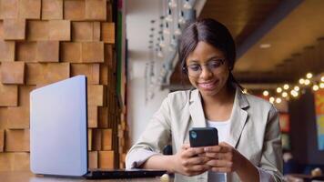 Young indian woman in glasses using phone while sitting in cafe with laptop video