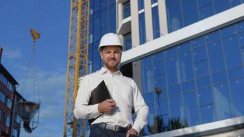 Architect engineer in a white shirt and helmet on a background of a modern glass building holds a tablet video