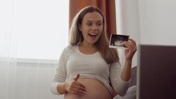 Pregnant fair-skinned woman shows the interlocutor a photo from the ultrasound examination of the fetus using communication on a laptop video