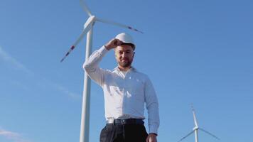 Air power engineer stands in business style clothes and white helmet on a background of windmill video