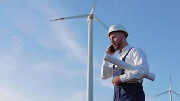 une Masculin ingénieur dans une blanc casque, travail bleu salopette et une classique chemise détient dans le sien main une grand roulé feuille de papier avec dessins de le projet et pourparlers sur le téléphone portable près le vent turbines video