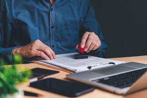 A person's hands are seen stamping a document, symbolizing the approval process in a professional setting. The laptop in the background suggests a modern office environment. photo