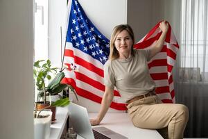Portrait of happy young woman with USA flag learning American English online from home using laptop. Smiling female student sitting at table and taking online educational course in foreign languages. photo