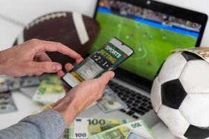 Man betting on sports using smartphone and laptop at table, closeup. Bookmaker websites on displays photo