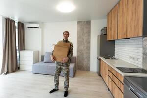 Smiling man in camouflage holding cardboard box in new apartments photo