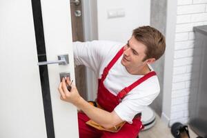 Installation of a lock on the front wooden entrance door. Portrait of young locksmith workman in blue uniform installing door knob. Professional repair service. Maintenance Concept photo