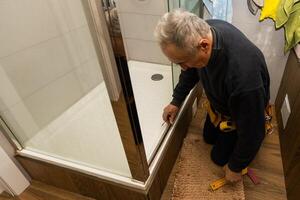 an elderly man repairing door of shower cabin in bathroom photo