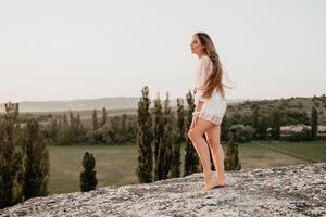 Happy woman in white boho dress on sunset in mountains. Romantic woman with long hair standing with her back on the sunset in nature in summer with open hands. Silhouette. Nature. Sunset. photo