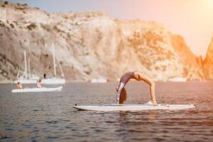 Woman sup yoga. Middle age sporty woman practising yoga pilates on paddle sup surfboard. Female stretching doing workout on sea water. Modern individual female hipster outdoor summer sport activity. photo