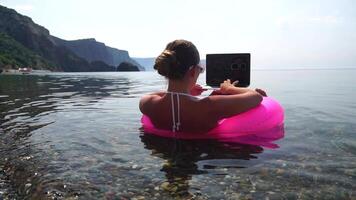 Woman freelancer works on laptop swimming in sea on pink inflatable ring. Happy tourist in sunglasses floating on inflatable donut and working on laptop computer in calm ocean. Remote working anywhere video