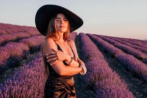 mujer lavanda campo. contento despreocupado mujer en negro vestir y sombrero con grande borde caminando en un lavanda campo durante puesta de sol. Perfecto para inspirador y calentar conceptos en viaje y pasión de viajar. foto