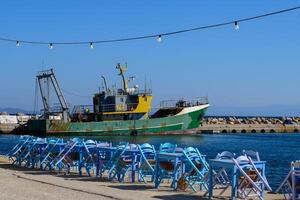 Blue wooden tables and chairs of a typical fish tavern at the port. Fishing ship on the background. Aegean island Chios in Greece on an autumn day. Greek holidays and destinations photo