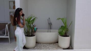 Indonesian woman wearing a pattern sleeveless top smiling while talking on phone call and walking at villa, working on holiday video