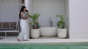 indonesiano donna indossare un' modello senza maniche superiore sorridente mentre parlando su Telefono chiamata e a piedi a villa, Lavorando su vacanza video
